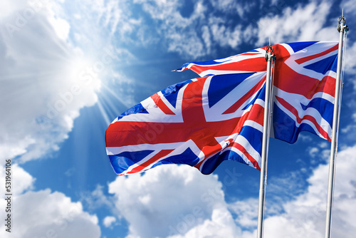 Two national UK flags with flagpole, blowing in the wind on a blue sky with clouds and copy space and sunbeams. Union Jack Flag.