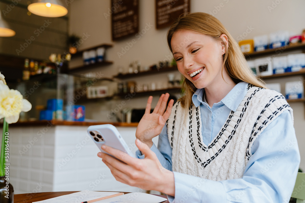 Blonde white woman waving hand and using mobile phone at cafe