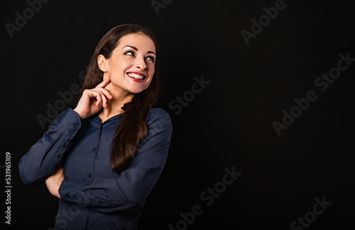 Beautiful thinking toothy smiling business woman looking up with folded arms in blue shirt on black background with empty copy space for text. Closeup