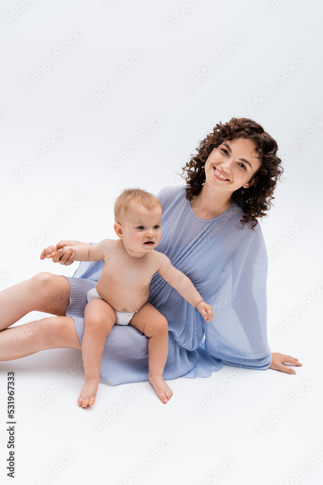 Mother in blue dress holding hand of infant daughter and looking at camera on white background.