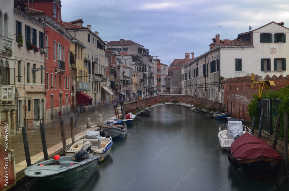 Long exposure of a Venetian street with boats on a canal in the foreground and a bridge in the background. People in motion are on the pavement.