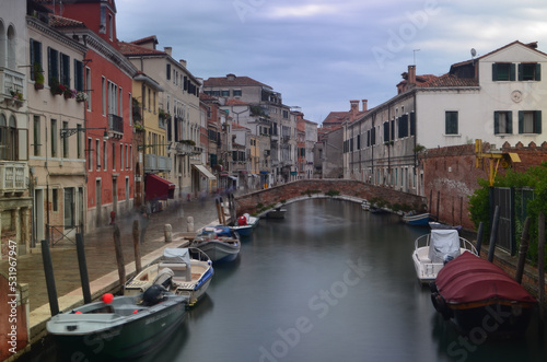 Long exposure of a Venetian street with boats on a canal in the foreground and a bridge in the background. People in motion are on the pavement. © Vojtch