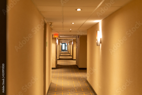 A long narrow hallway of a hotel with a red exit sign light sconce lights and cream colored walls. The carpet on the floor is green and beige with a pattern. The low ceiling has white tiles. 