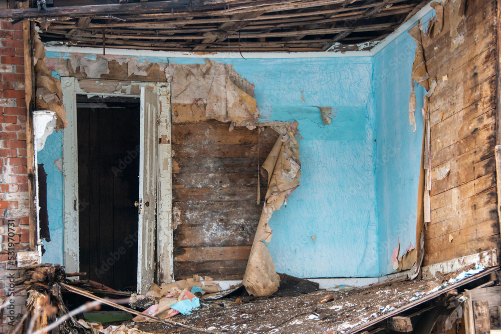 The interior of a building being demolished. The walls are layers of blue paint, wallpaper, and wood. There's an open single white wooden door in the house. The room is covered in dirt and materials. The interior of a building being demolished. The walls are layers of blue paint, wallpaper, and wood. There's an open single white wooden door in the house. The room is covered in dirt and materials.