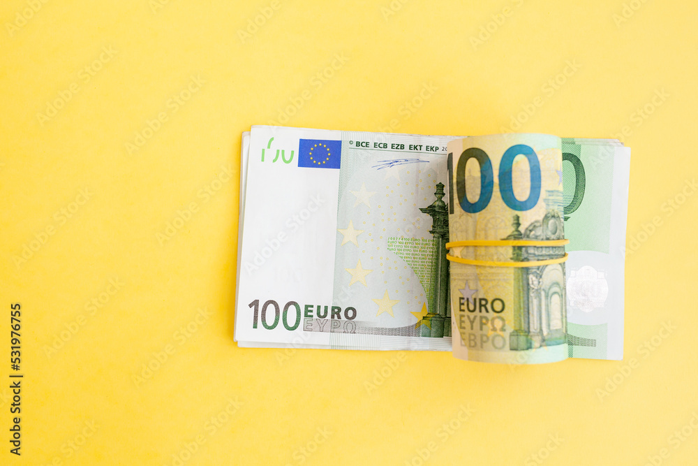 Money roll of euro banknotes with a elastic band on a 100 euro paper bill on a yellow background. cash currency, payment, earnings and savings, the concept of money and finance. Empty space for text Money roll of euro banknotes with a elastic band on a 100 euro paper bill on a yellow background. cash currency, payment, earnings and savings, the concept of money and finance. Empty space for text