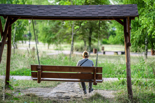 A rear view of a woman in a hat sitting on a wooden swing and looking ahead in a suburban park. Rest against the background of nature. The concept of a summer lifestyle.