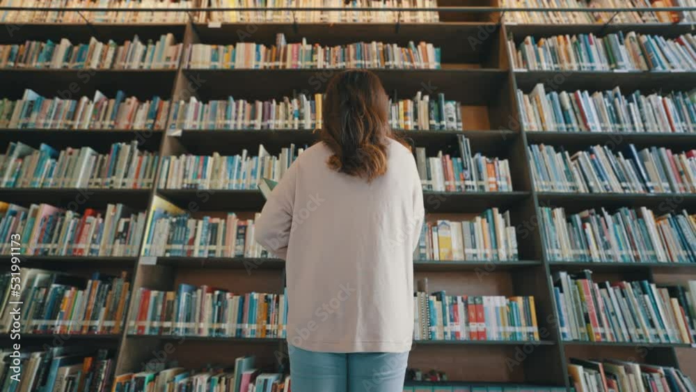 Back view of a student looking at rows of bookshelves searching for class assignments and exam preparations in the university library. Back view of a student looking at rows of bookshelves searching for class assignments and exam preparations in the university library.