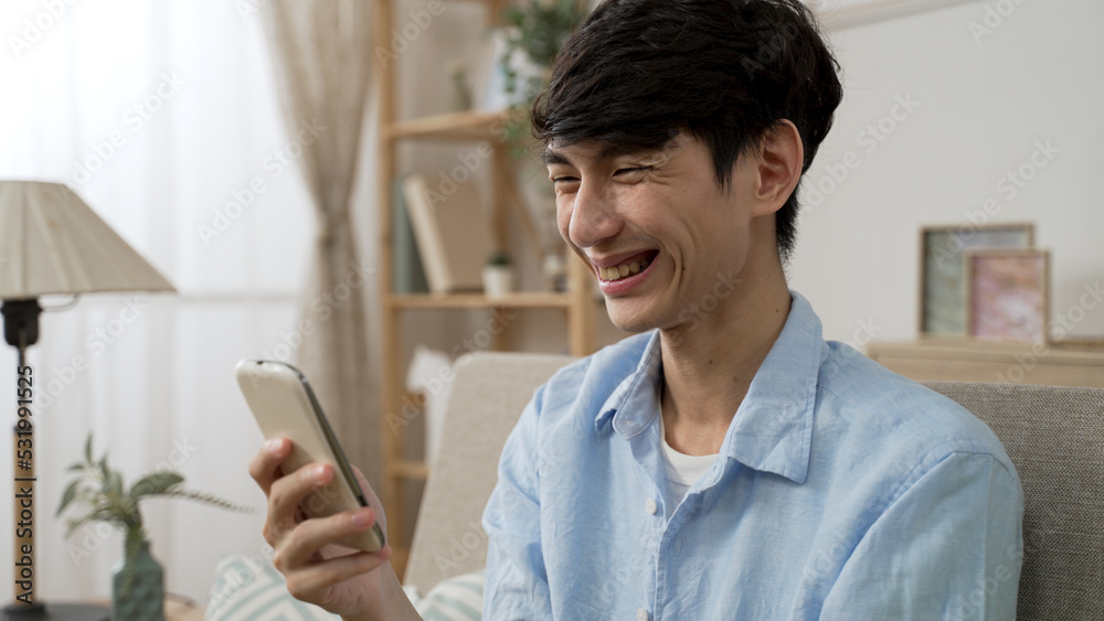 closeup of a carefree asian male laughing at a funny post on social media website while playing with his mobile phone in the living room at home closeup of a carefree asian male laughing at a funny post on social media website while playing with his mobile phone in the living room at home