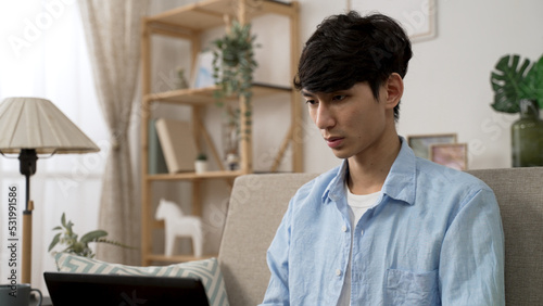 concentrated asian Japanese man frowning with a thoughtful face while working from home on the computer in the living room at daytime