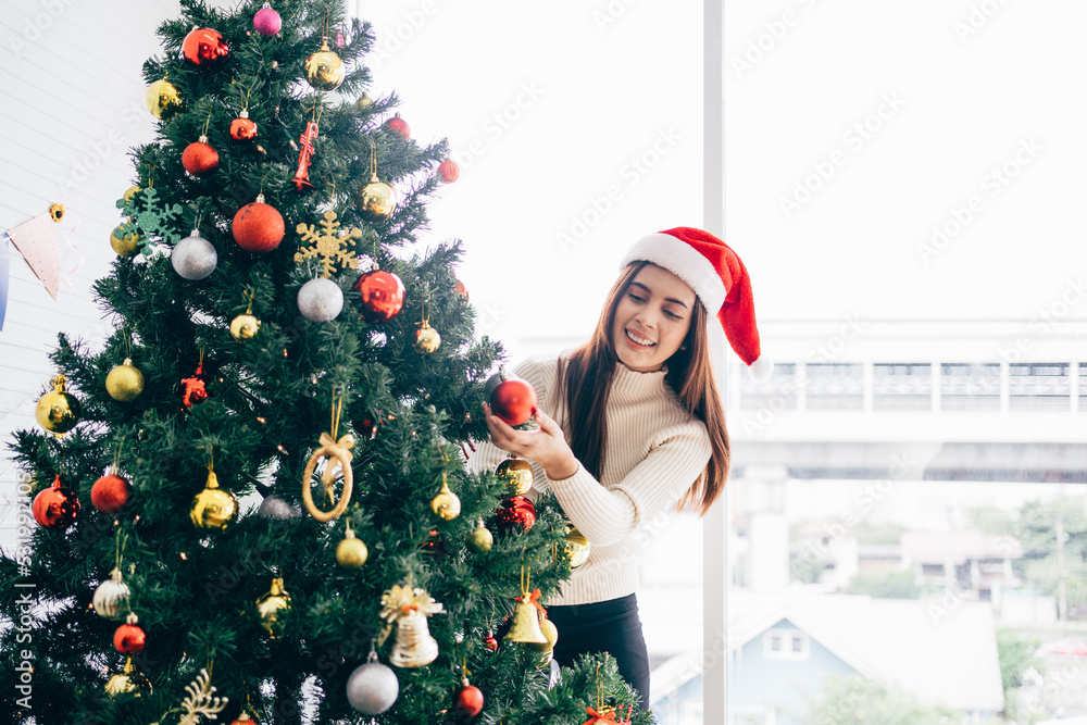 Happy young woman wearing a Santa Claus hat and sweater, smile and enjoy decorating various baubles on the Christmas tree in the living room at home. Enjoying and celebrating the Christmas holiday. Happy young woman wearing a Santa Claus hat and sweater, smile and enjoy decorating various baubles on the Christmas tree in the living room at home. Enjoying and celebrating the Christmas holiday.