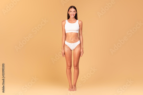 Full length frontal view of slim woman with perfect body shape and flat tummy posing on beige studio background