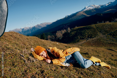 Woman lying on a hill on the grass rest smile with teeth looking at the mountains in the snow in winter in a yellow raincoat and jeans happy sunset trip on a hike, freedom lifestyle 