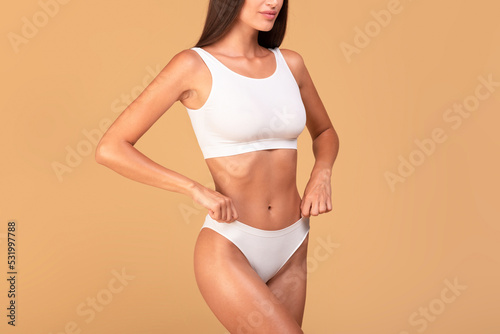 Unrecognizable well-fit lady in white underwear demonstrating her perfect body, posing over beige studio background