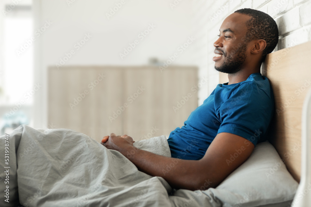 Cheerful Black Guy Sitting In Bed In Bedroom, Side View Cheerful Black Guy Sitting In Bed In Bedroom, Side View