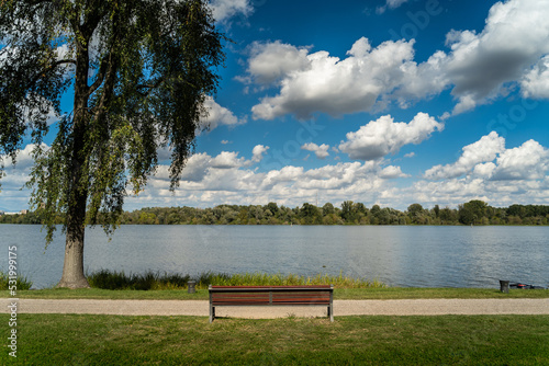 A wonderful sunny day walking along the Mincio river in Mantua, Italy. A lots of clouds dominate the sky together with the green of the meadows and a lonely bench.