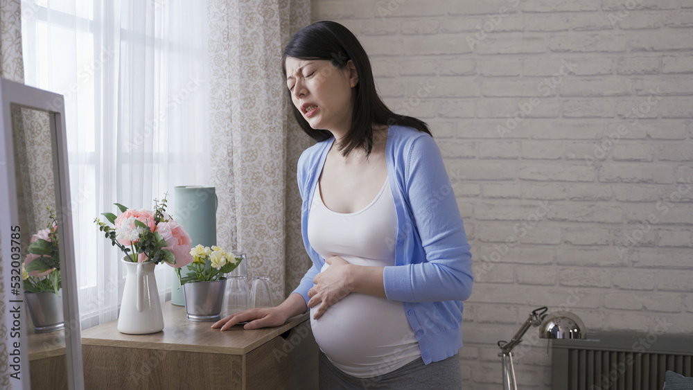 waist up japanese expectant mother propping herself on the cabinet is suffering labor contraction with a painful expression at home. waist up japanese expectant mother propping herself on the cabinet is suffering labor contraction with a painful expression at home.