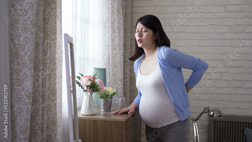 asian woman approaching to the window is massaging herself while feeling sudden lower back pain during pregnancy in the morning at home.