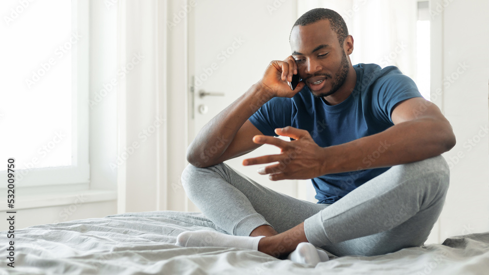 Black Male Talking On Mobile Phone Sitting In Modern Bedroom