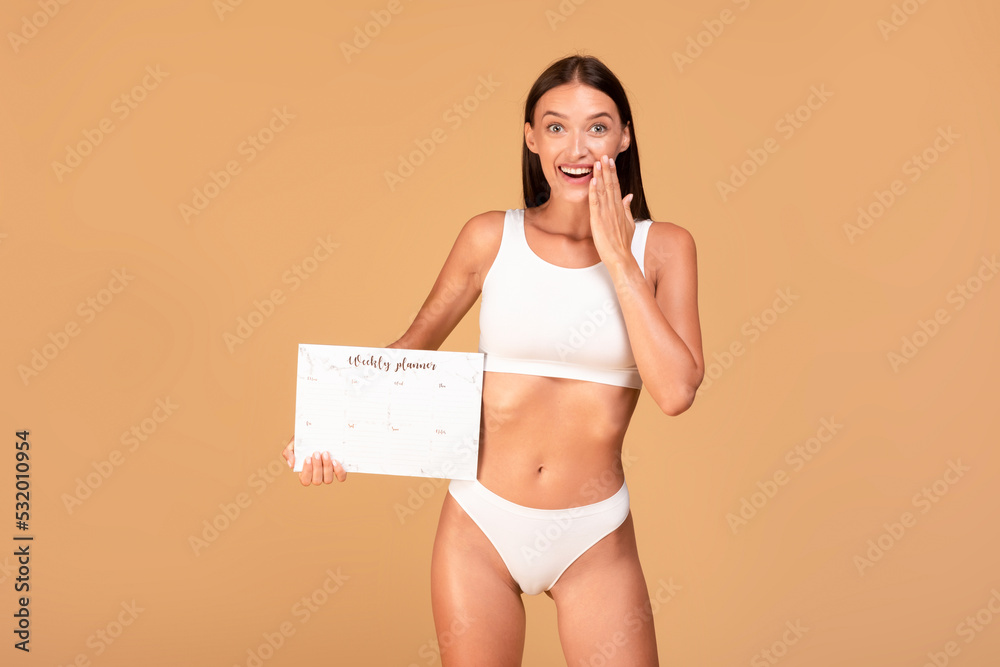 Fototapeta premium Excited fit woman in white underwear holding paper weekly planner, being happy of planning her diet and trainings