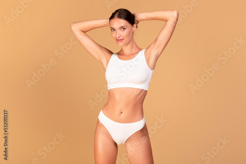 Portrait of happy young fit woman wearing lingerie posing over beige studio background, raising arms up