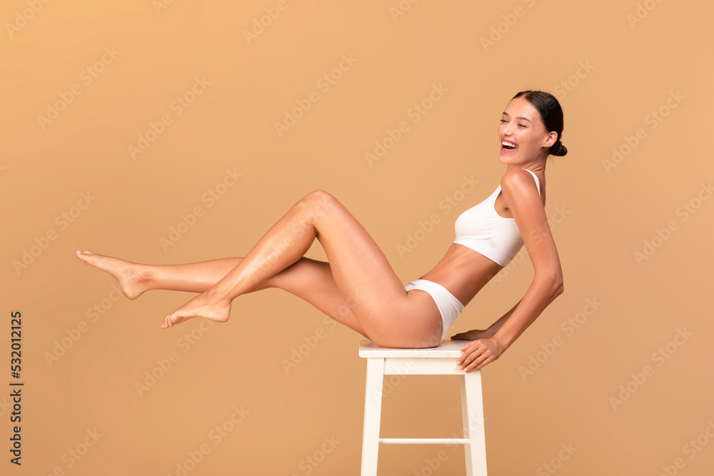 Overjoyed slim woman in white underwear sitting on white chair and smiling over beige studio background