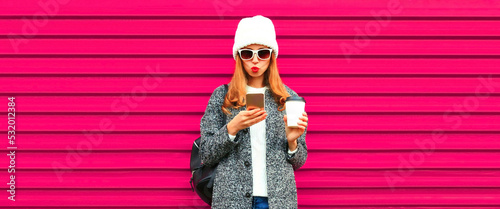 Portrait of stylish smiling woman with smartphone wearing white knitted hat, gray coat jacket on pink background © rohappy