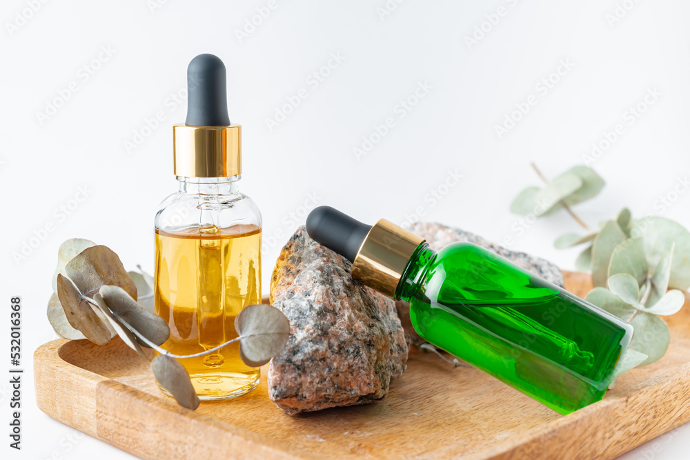 Naklejka premium glass dropper bottles with a pippette with gold rubber tip and green eucalyptus leaves near it on wooden tray on white background. Nature Skin concept. Organic Spa Cosmetics.
