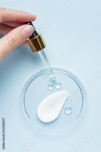 Female hand holding a pipette dropper with face beauty oil over petri dish with face cream strokes. Laboratory research. Herbal cosmetics