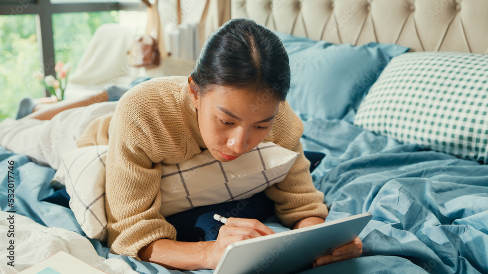 Happy Asian girl university with white cream pajamas lay down on fluffy comfort bed use digital tablet learning online in bedroom at home. Revisualizing flexible working, Work from home concept. Happy Asian girl university with white cream pajamas lay down on fluffy comfort bed use digital tablet learning online in bedroom at home. Revisualizing flexible working, Work from home concept.