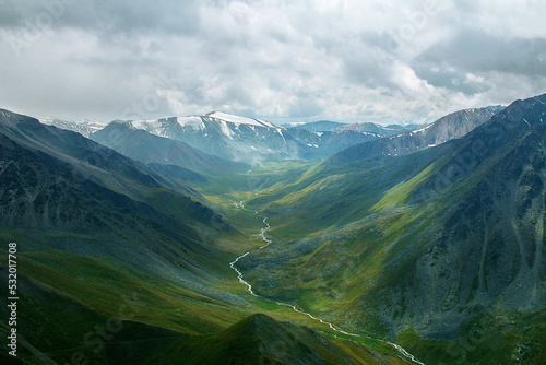 A beautiful gorge with high green slopes, a river flows along the gorge, the tops of the mountains are covered with snow, the sky is in thick clouds, summer is in the mountains