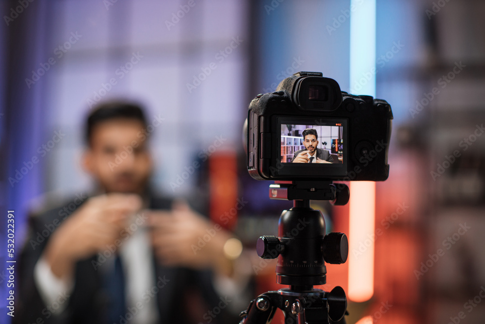 View from camera screen of confident indian bearded businessman, broker in suit sitting in front of camera in evening office during recording video for business vlog.