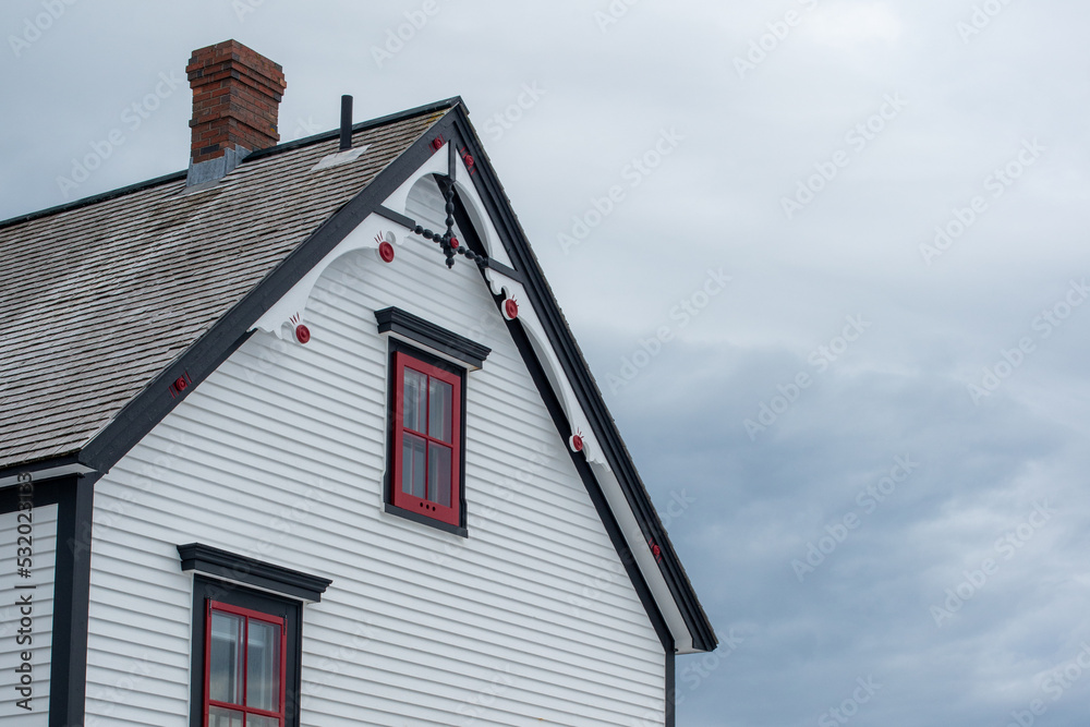 Exterior white wooden wall of horizontal clapboard siding on a vintage house with black trim. There are small four pane glass windows with red trim and a decorative gable cedar shake roof.