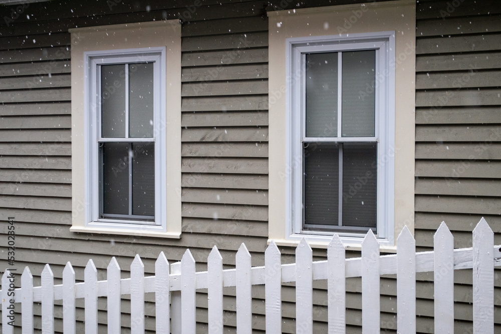Fototapeta premium White picket fence on a diagonal in the foreground with a tan coloured house in the background. There are two white double hung windows with beige trim on the exterior wall of the country style home.