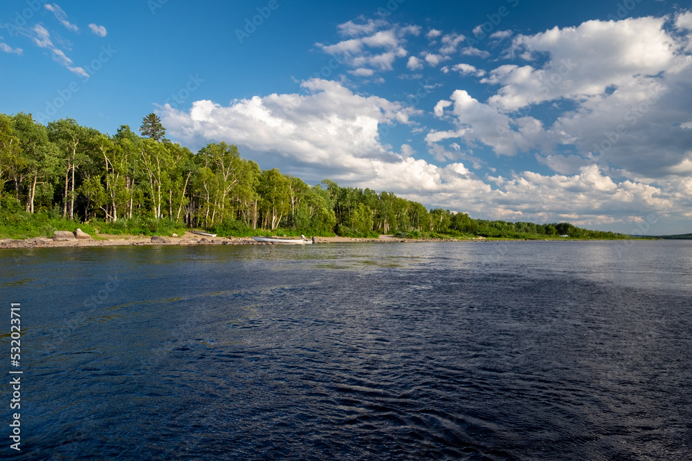 A wide natural river with smooth blue water. There are luscious green trees along the riverbank. The sky is clear blue. The calm and peaceful pond water is reflecting the sky and tall green trees.