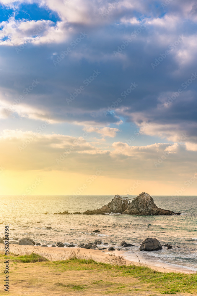 Sunset sky above the lawn on the coast bordered by wet rocks along the Itoshima Beach of Fukuoka with the Sakurai Futamigaura's Meoto Iwa Couple Rocks protected by a sacred Shinto straw rope.