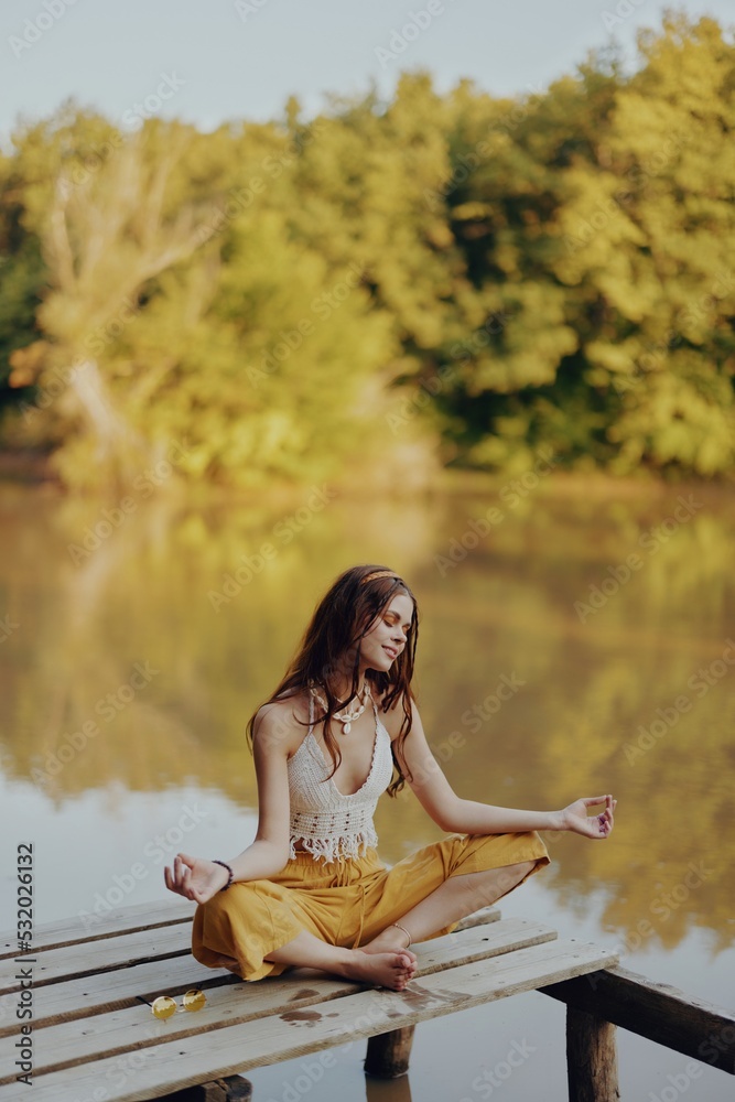 A young happy woman with a hippie smile sits on the lake shore on a bridge wearing eco clothing made of natural materials in harmony with nature in the fall. Travel lifestyle
