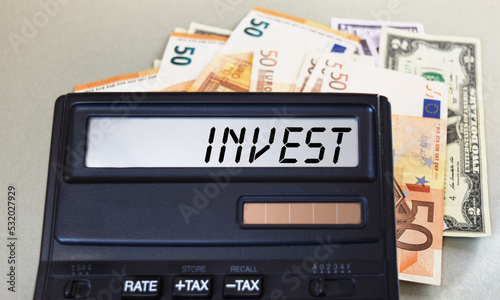 INVEST text written on a calculator next to the background of euro banknotes. Finance or business concept.