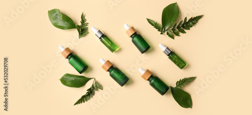 Different bottles of cosmetic serum and green leaves on beige background, top view