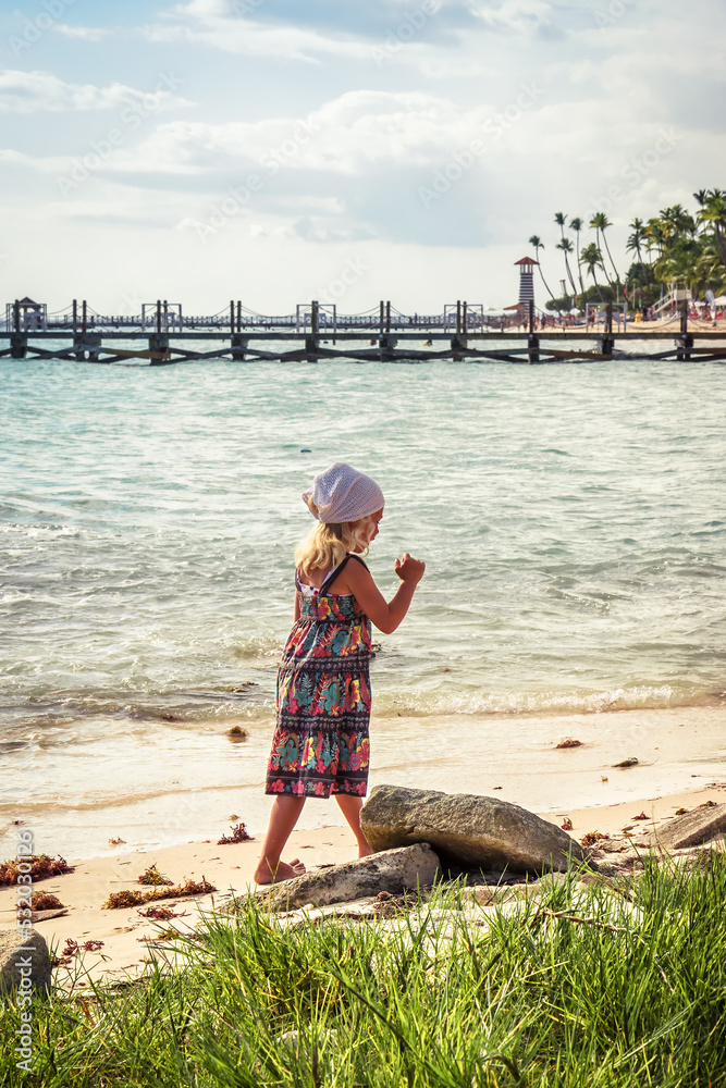 Small child girl walking alone on tropical beach with sea pier and lighthouse on background as summer vacation lifestyle