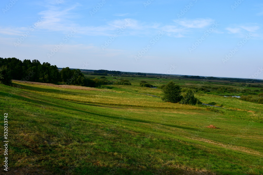 A view from the top of a tall hill with numerous trees, shrubs, and forest formations visible next to some fields, meadows, and pasturelands spotted on Polish countryside in summer
