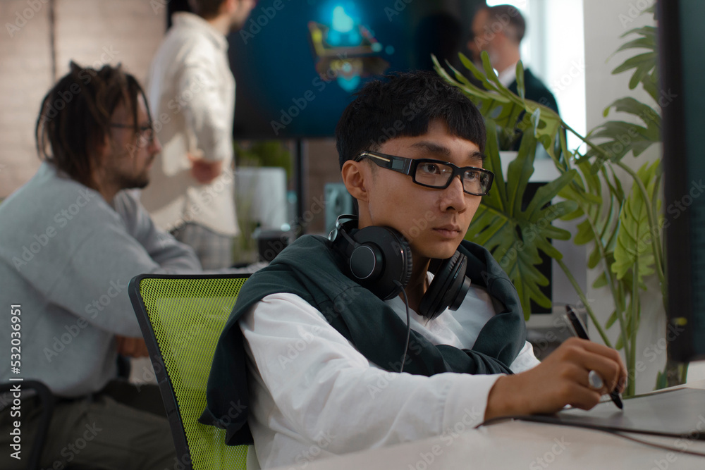 Asian male artist works in headphones in a studio for the development of computer games and applications. In the background, a team of developers are discussing a 3D model. Asian male artist works in headphones in a studio for the development of computer games and applications. In the background, a team of developers are discussing a 3D model.