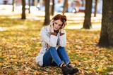 Unhappy sad caucasian millennial red-haired woman in raincoat thinks, sits on ground in park with yellow leaves