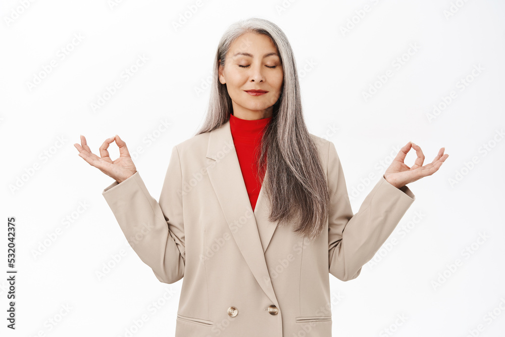 Keep calm. Asian senior woman in business suit, meditating, deep breath, inhale air and relaxing, standing over white background Keep calm. Asian senior woman in business suit, meditating, deep breath, inhale air and relaxing, standing over white background