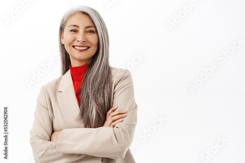 Portrait of successful asian senior businesswoman, smiling and looking happy, professional power pose, corporate outfit, standing over white background