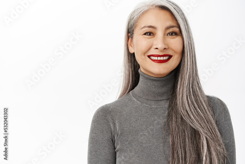 Aging and cosmetology concept. Portrait of beautiful and healthy middle aged asian woman with makeup, grey long hair, smiling and looking happy, white background