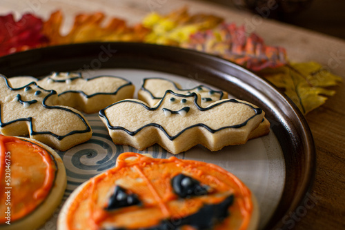 Halloween Bat Sugar Cookies and Pumpkin Jack-O-Lantern Cookies on Plate with Fall and Autumn Leaves, Sitting on Dark Wood Table. 