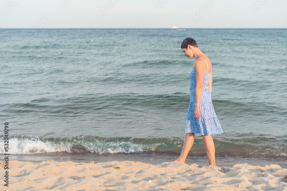 Attractive short haired woman in her thirties wear in light summer dress walks barefoot on the beach with sea in the background. Concept for vacation, leisure, relaxation, physical and mental health. Attractive short haired woman in her thirties wear in light summer dress walks barefoot on the beach with sea in the background. Concept for vacation, leisure, relaxation, physical and mental health.