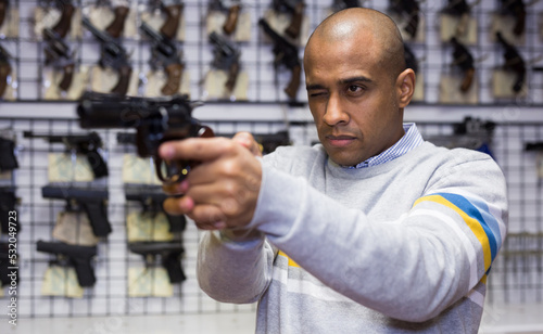 Focused latin american man examining convenience and accuracy of new pistol in gun shop, imitating aiming