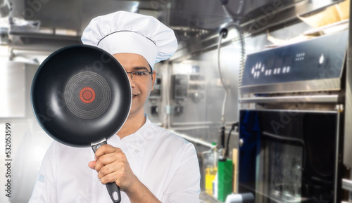Chef hides face behind frying pan. Male cook in white uniform. Cook is trying to hide. Man chef with frying pan in front of his face. Guy cook is standing in kitchen. Restaurant employee smiling