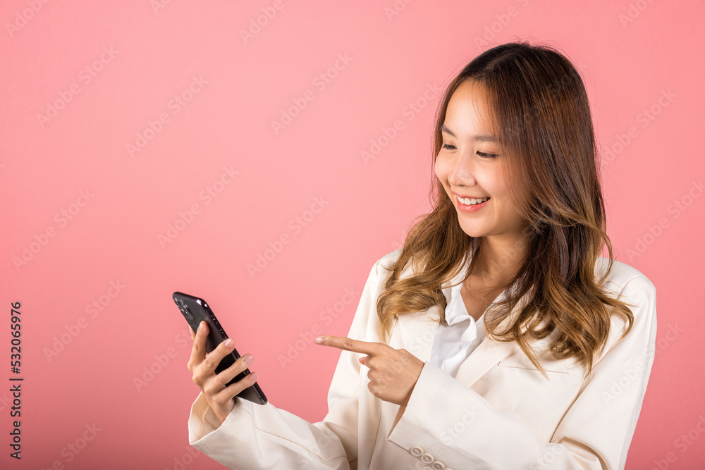 Portrait Asian happy beautiful young woman smile making finger pointing on screen smartphone in hand shopping online application looking to screen, studio shot isolated pink background with copy space Portrait Asian happy beautiful young woman smile making finger pointing on screen smartphone in hand shopping online application looking to screen, studio shot isolated pink background with copy space